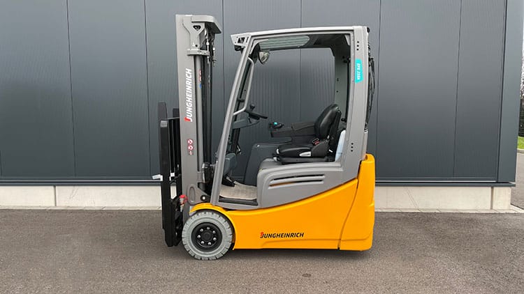 Gelber und grauer Jungheinrich Gabelstapler steht auf Asphalt vor einer dunkelgrauen Industriehalle.