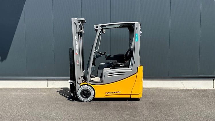 Gelber und grauer Jungheinrich Gabelstapler steht auf Asphalt vor dunkler Industriehallenwand.