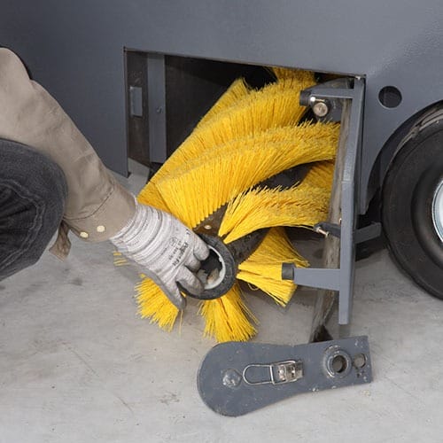Eine Person mit Handschuhen montiert oder entfernt eine gelbe Zylinderbürste aus einer Bodenreinigungsmaschine.