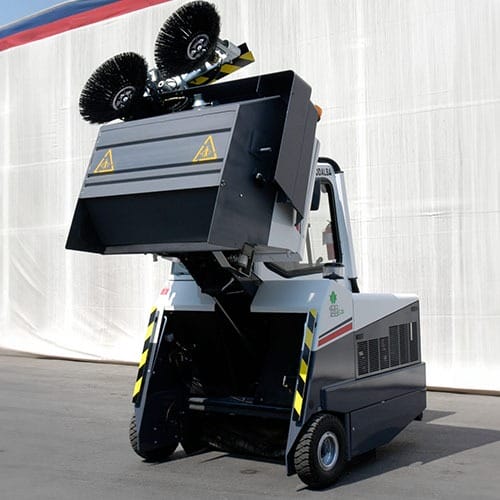 Kehrmaschine mit angehobenen rotierenden Bürsten und offenem Sammelbehälter steht auf Asphalt vor weißer Stoffwand.