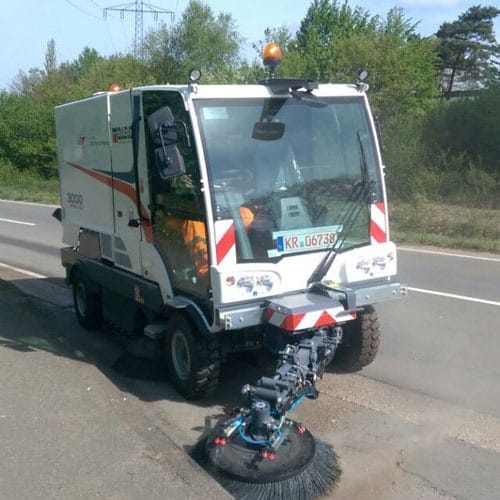 Kehrmaschine reinigt Straßenrand mit rotierender Bürste; Bäume und Straßenschutzplanke im Hintergrund sichtbar.