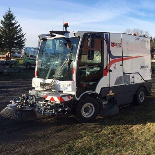 Straßenreinigungsfahrzeug mit rotierenden Bürsten steht tagsüber auf gepflasterter Fläche nahe Rasen und Bäumen im Freien.