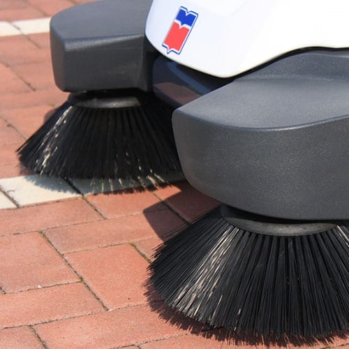 Nahaufnahme von zwei rotierenden Bürsten einer Bodenreinigungsmaschine fegen Pflastersteine auf einem Gehweg im Freien.