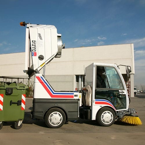 Kompakte Kehrmaschine mit hochgeklappter Mulde steht neben grünem Müllcontainer vor großer Industriehalle.
