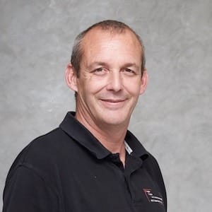 Mann mit kurzem grauem Haar lächelt, trägt dunkles Hemd vor hellem Hintergrund; Porträt für Technische Industrieberatung.