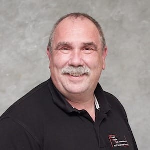 Mann mit kurzem grauem Haar lächelt, trägt dunkles Hemd vor hellem Hintergrund; Porträt für Technische Industrieberatung.