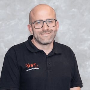 Mann mit kurzem grauem Haar lächelt, trägt dunkles Hemd vor hellem Hintergrund; Porträt für Technische Industrieberatung.