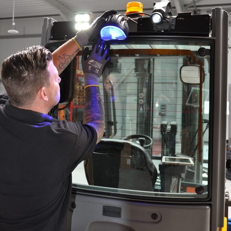 Ein Mann mit Handschuhen prüft mit Blaulichtgerät die Frontscheibe eines Gabelstaplers in einer Lagerhalle oder Werkstatt.